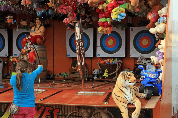 A little girl standing in front of a display of stuffed animals. Year market bude archery.